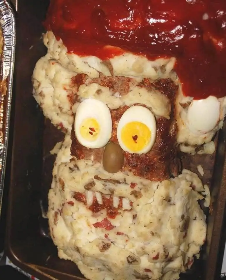 A meatloaf dish creatively shaped and decorated to resemble a face, with halved hard-boiled eggs as eyes, a green olive as a…