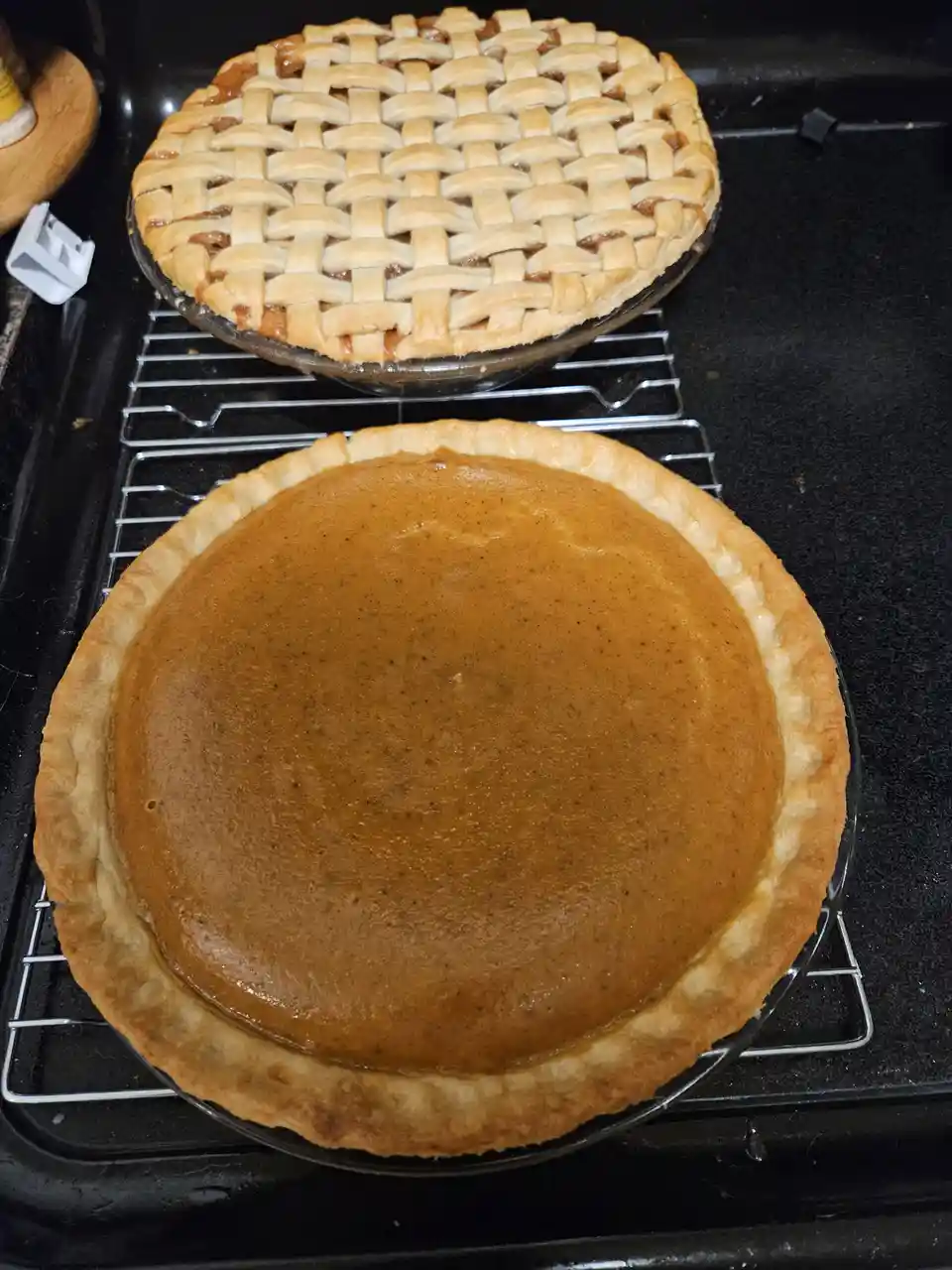 Two freshly baked pies sit on cooling racks on a stovetop: a pumpkin pie with a smooth, golden-brown filling and a plain…