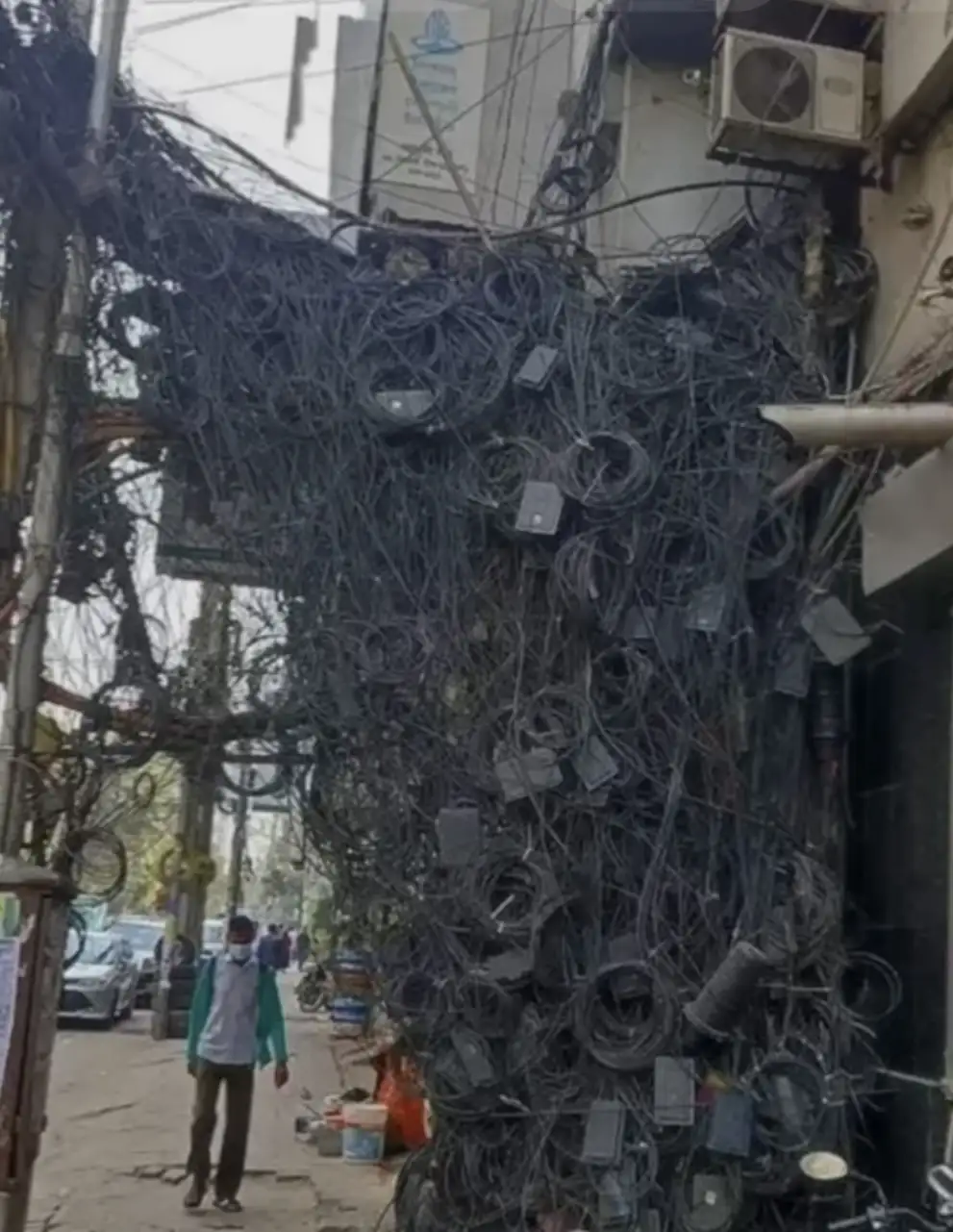 A street scene shows a massive, chaotic tangle of black electrical and communication wires draped over a building and…