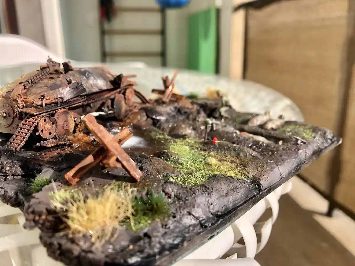 A close-up photo shows a detailed, weathered model of a tank wrecked and stuck in a muddy, grassy diorama. The tank is…