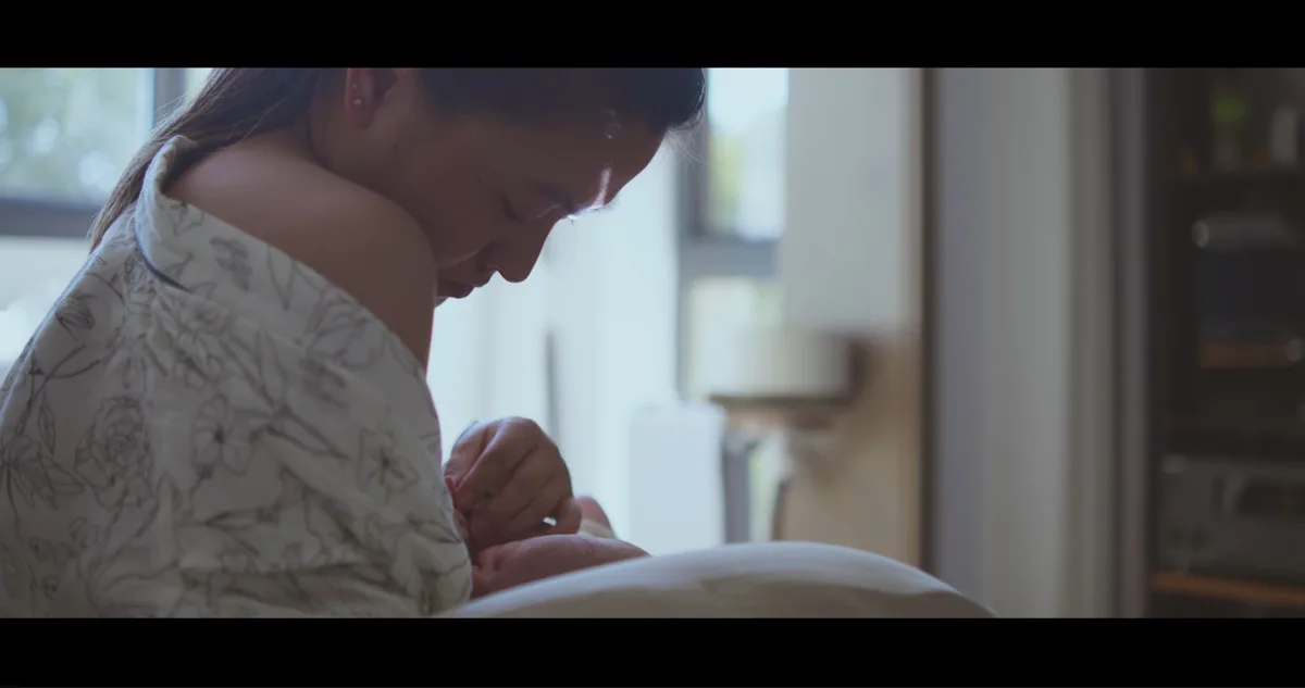 A woman in a floral top leans over a baby in a crib, gently touching the infant's hand. The image has black bars on top and…