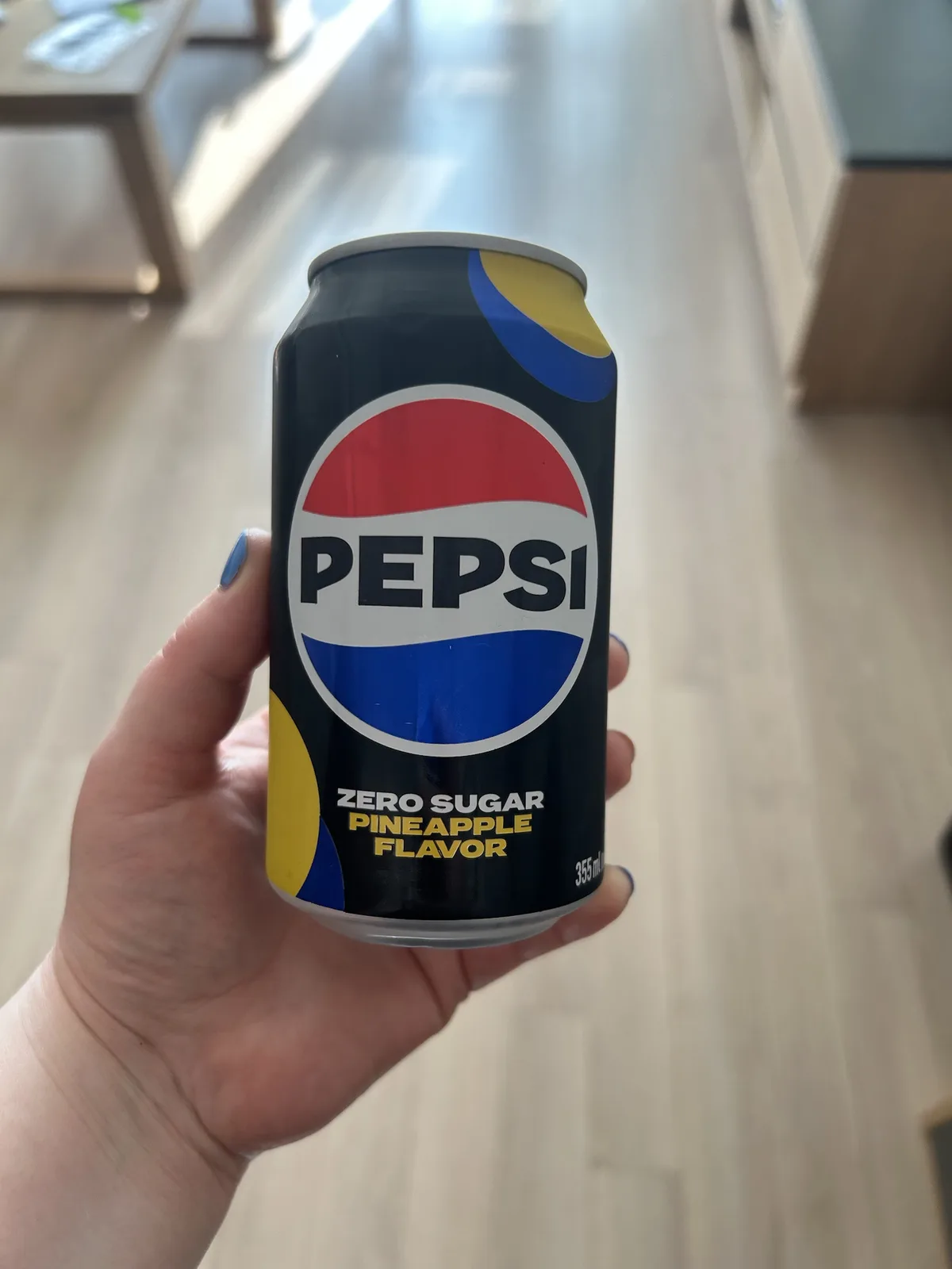 A hand with blue nail polish holds a black Pepsi can labeled 'ZERO SUGAR PINEAPPLE FLAVOR' against a blurred wooden floor…