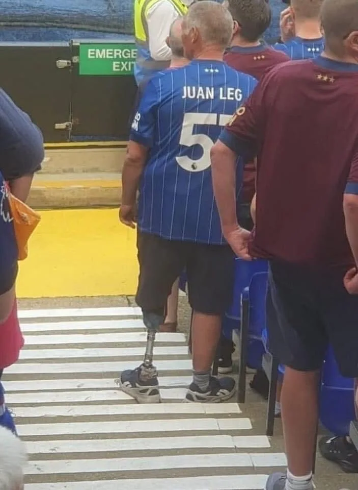 A man with a prosthetic leg stands on a zebra crossing, wearing a blue football jersey with 'JUAN LEG' and the number 5 on…