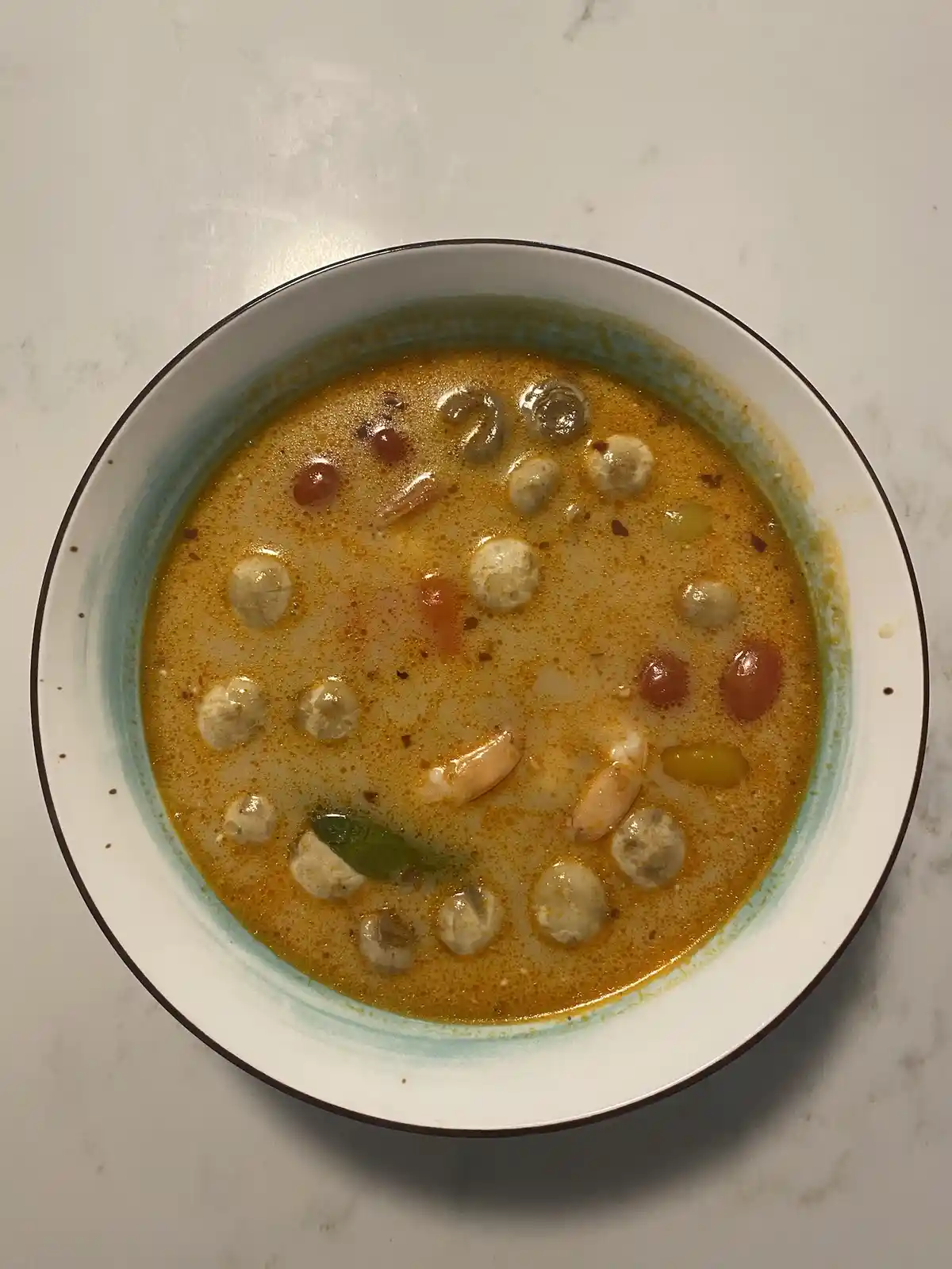 A top-down photo of a bowl of orange-colored curry soup with visible ingredients like shrimp, small round dumplings, cherry…