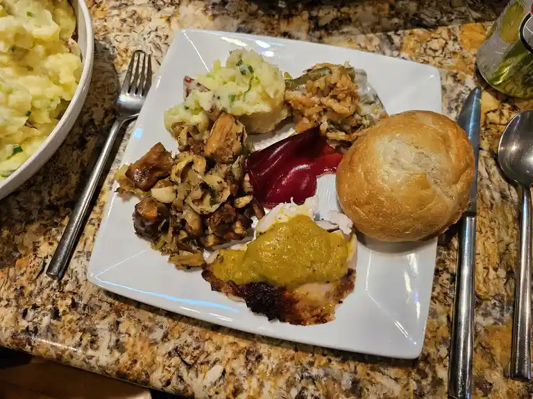 A white square plate holds a large, messy serving of food including mashed potatoes, green bean casserole, a bread roll, a…