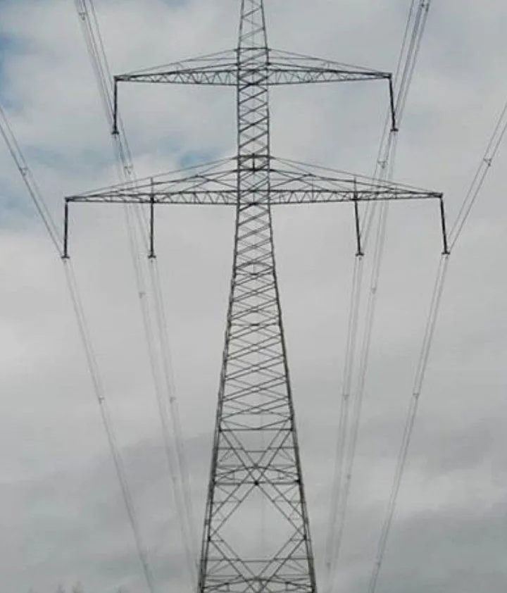 A tall, metal lattice electricity pylon stands against a cloudy sky, with multiple power lines extending from its arms…