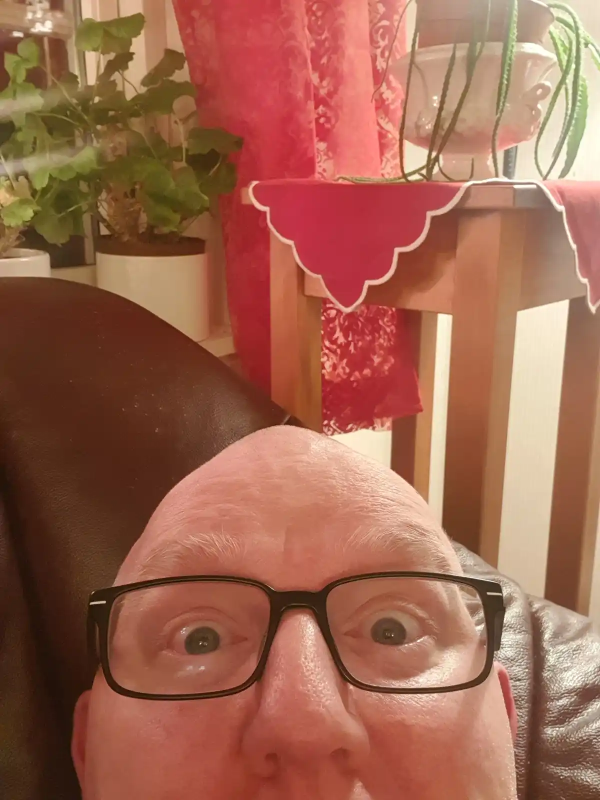 A close-up selfie of a bald man with black-rimmed glasses, his eyes wide open in a startled or comedic expression, seated in…