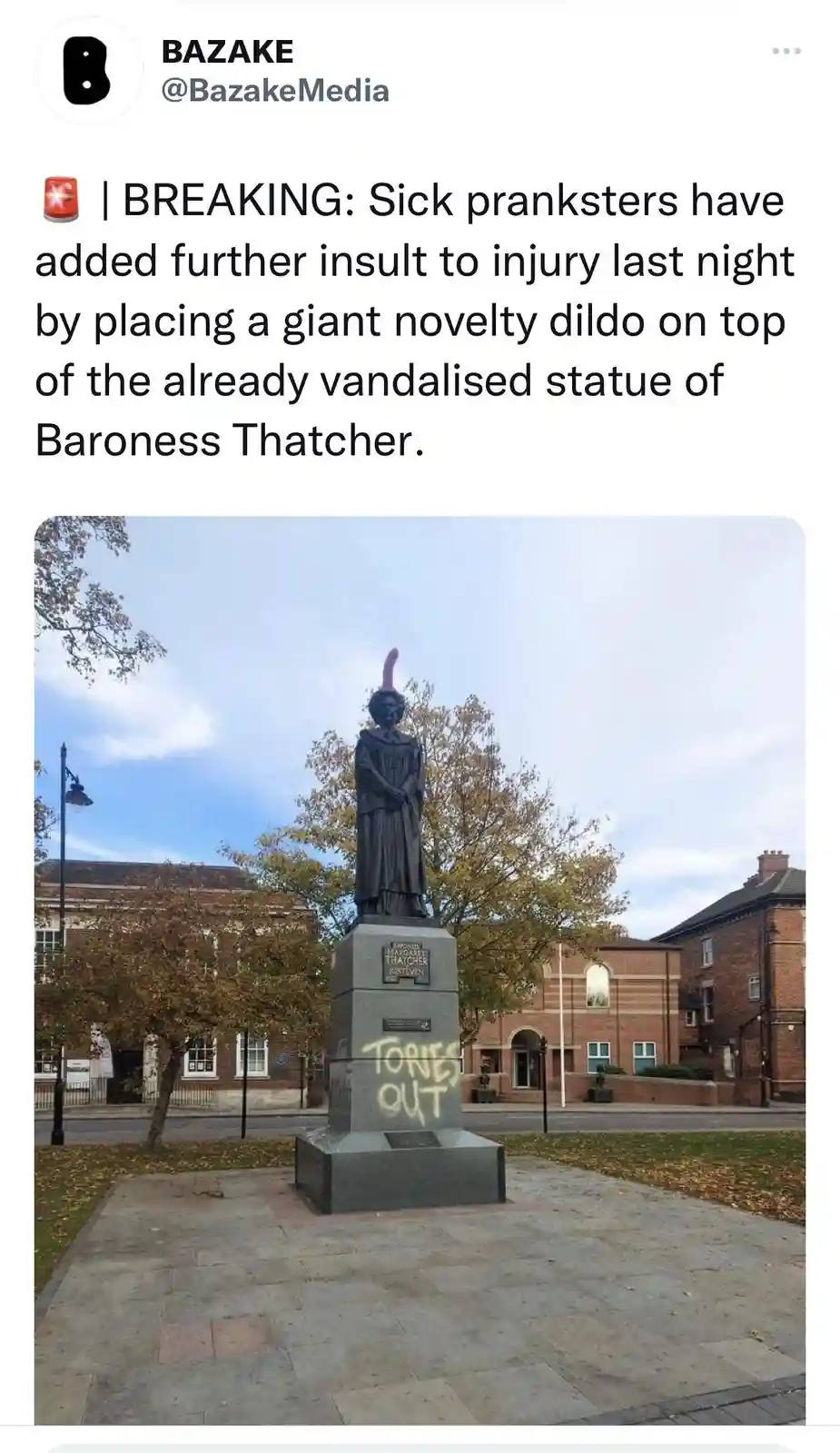 A photograph shows a dark statue of Baroness Thatcher on a stone pedestal in a public square, with trees and brick buildings…