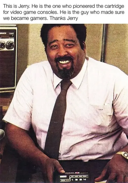 A smiling Black man with a goatee, wearing a white shirt and dark tie, sits at a desk holding a video game cartridge. Behind…