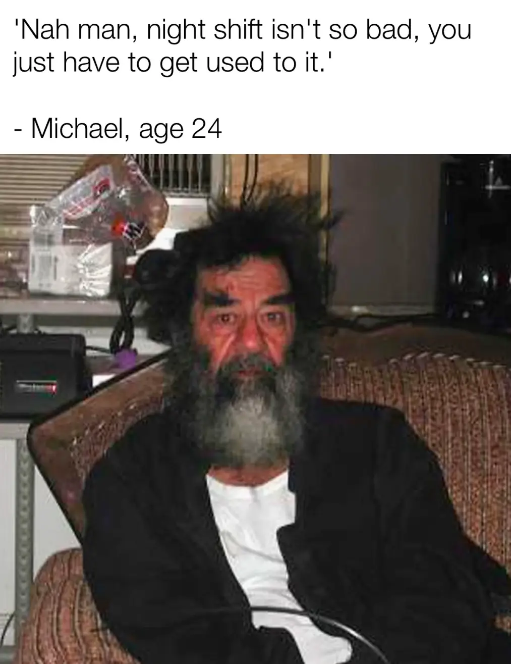 A man with wild, unkempt hair and a long, grizzled beard sits on a patterned couch looking tired and disheveled. The…