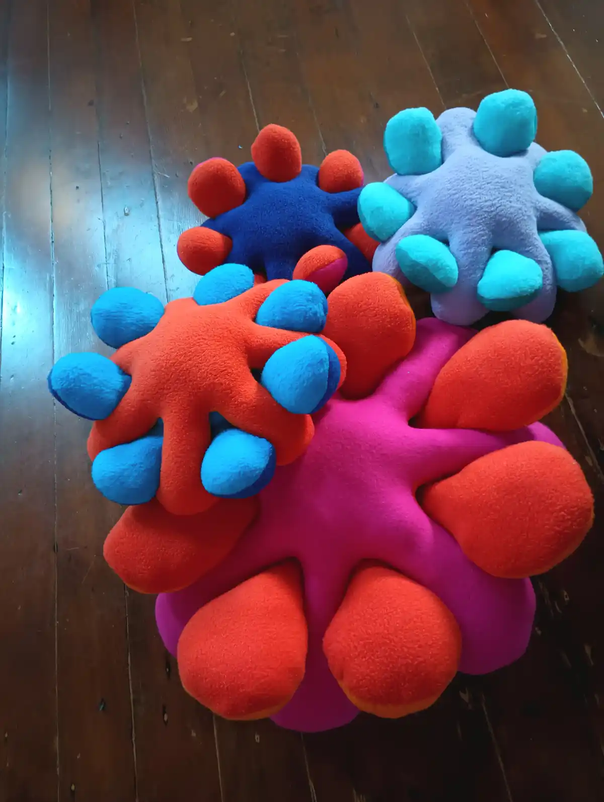 Four colorful, plush, virus-shaped toys with fuzzy textures and protruding bumps lie stacked on a wooden floor. The toys are…