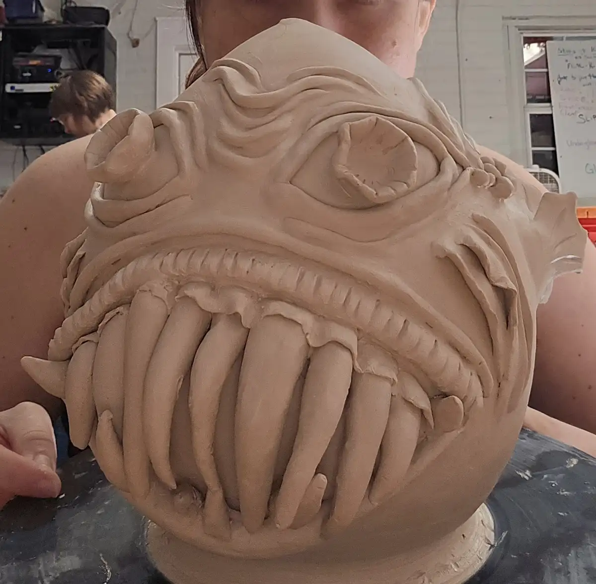 A person is holding a large, unfinished clay sculpture of a monstrous face with exaggerated features, including a wide mouth…