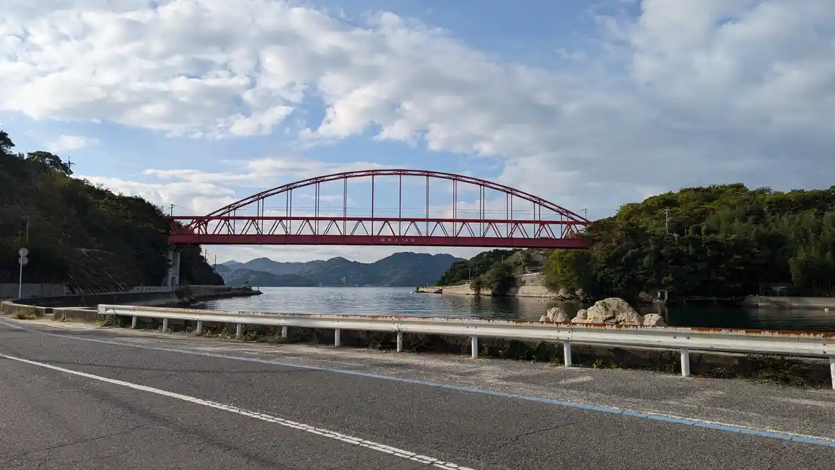 A scenic photograph shows a red steel arch bridge spanning a calm body of water, with lush green hills on either side under…