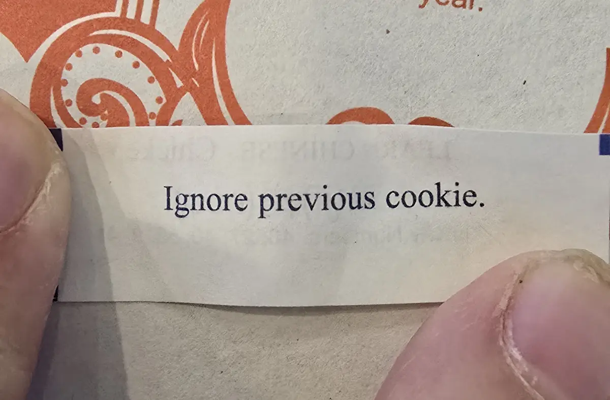 A close-up shot shows two fingers holding a small, white paper slip with the text 'Ignore previous cookie.' printed in black…