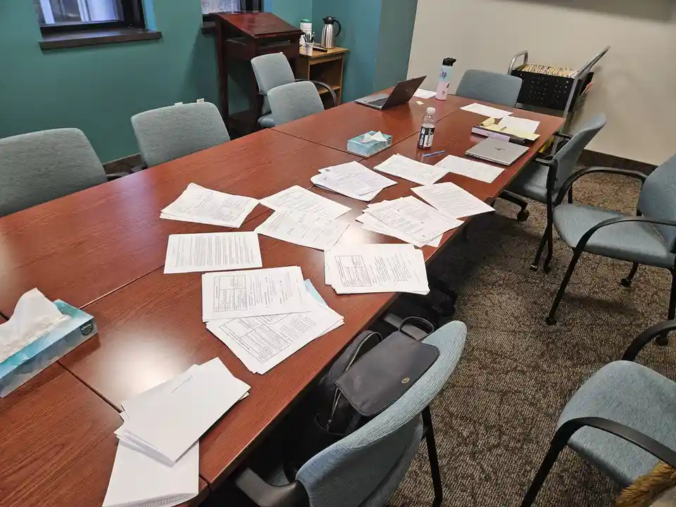 A conference room with a long wooden table covered in scattered papers, empty chairs, a laptop, a water bottle, and a tissue…