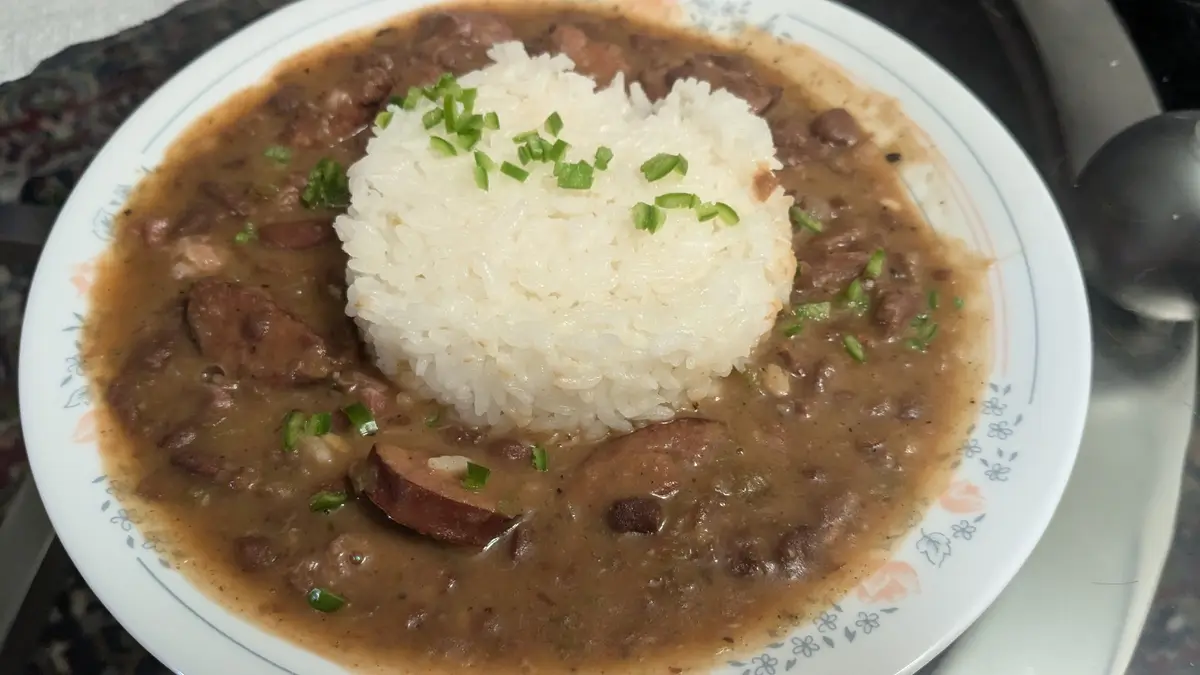A close-up photo shows a white plate with a mound of white rice topped with chopped green onions, surrounded by a brown stew…