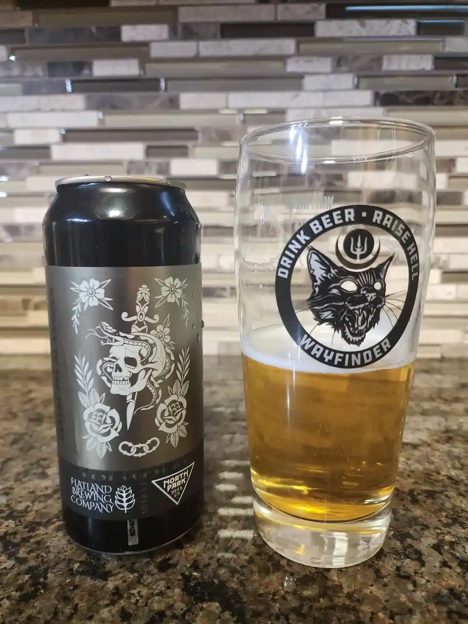 A black beer can with a skull and floral design sits next to a pint glass half-filled with beer. The glass features a…