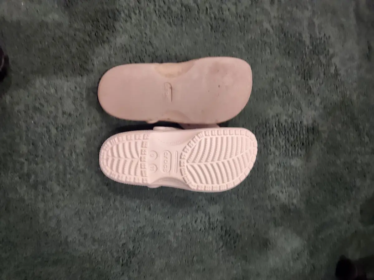 A top-down photo shows two light pink Crocs clogs on a dark green carpet, one shoe's sole is visible with the brand name and…