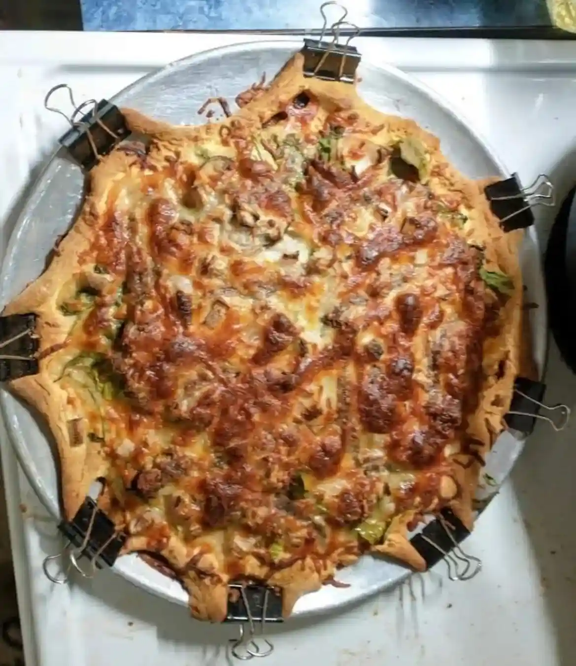 A cooked pizza with a distinctly irregular, star-like shape sits on a metal baking tray, held in place by multiple black…