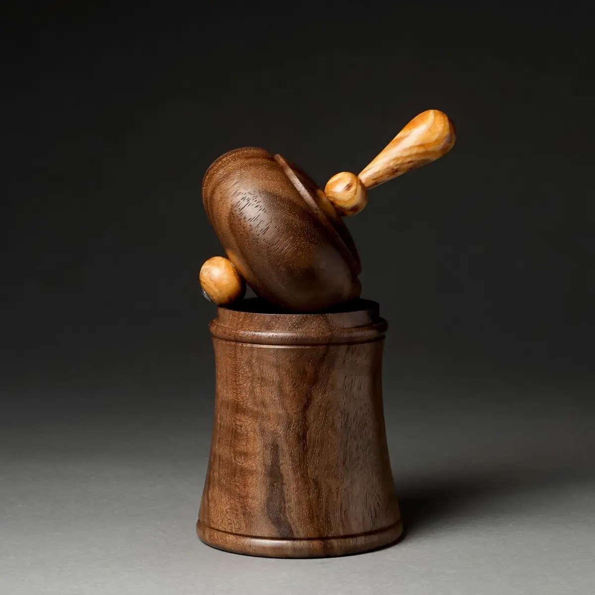 A wooden pepper mill with a handle is shown leaning precariously out of its wooden base against a dark background. The…