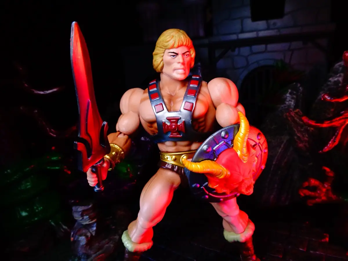 A He-Man action figure stands in a heroic pose, holding a red sword and a shield with a demonic face, against a dark…