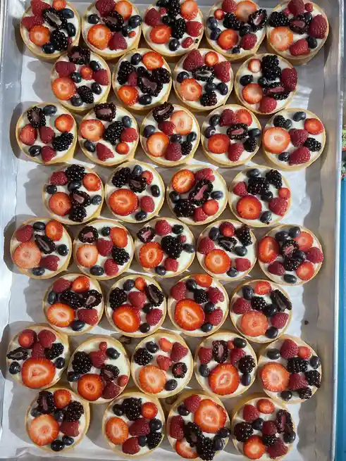 An overhead photo shows a metal baking tray lined with parchment paper, holding dozens of small, round fruit tarts. Each…