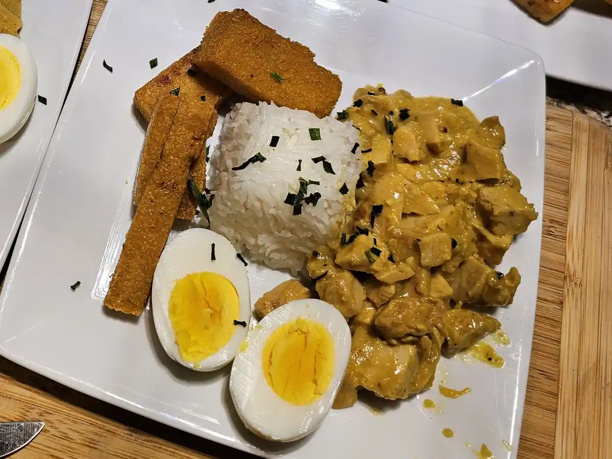 A white plate holds a meal of curry chicken, white rice, fried tofu, and two halves of a hard-boiled egg, all garnished with…