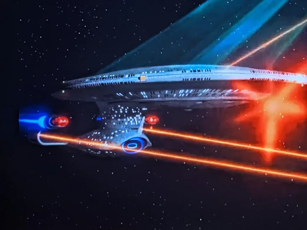 A Star Trek starship, resembling the Enterprise, is shown in space with blue and orange energy beams firing from its…