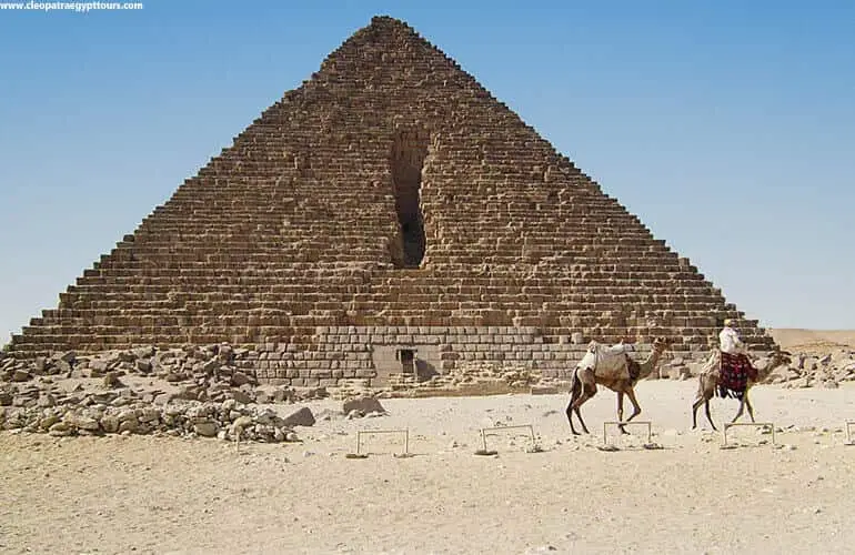 A photo of two camels with riders walking in front of a large pyramid in Egypt, with small hurdles placed on the sand…