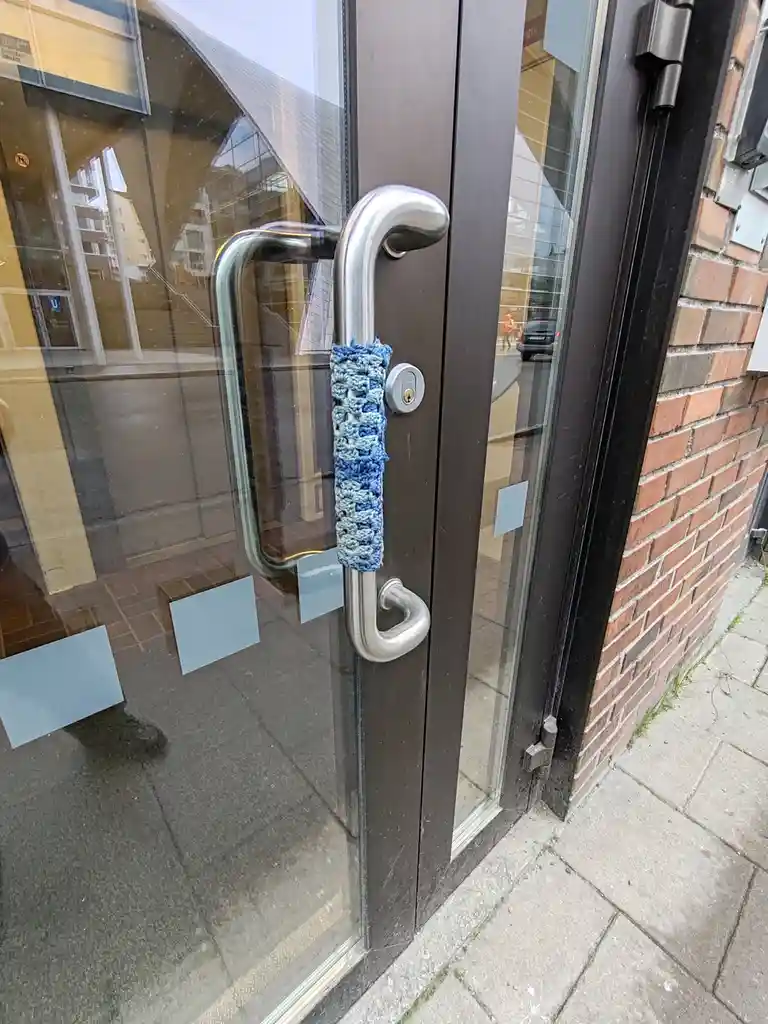 A close-up photo of a modern glass door with a silver metal handle wrapped in a blue and light blue knitted or crocheted…