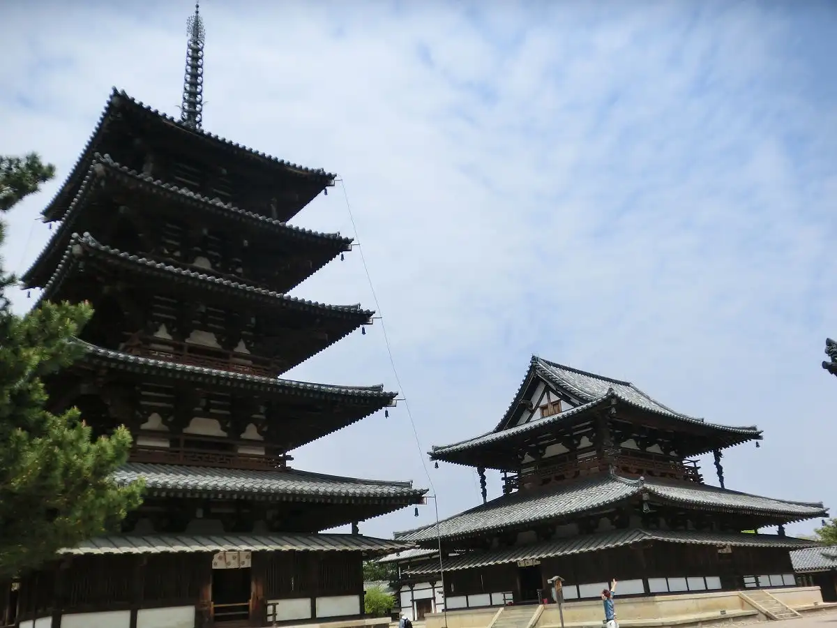 A low-angle photo shows a large, traditional Japanese pagoda and temple buildings under a partly cloudy sky. A person in a…
