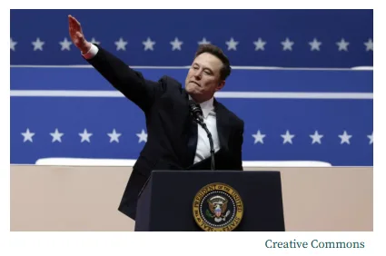 A photo of Elon Musk standing at a podium with the Presidential Seal, waving his right hand. The background is a blue wall…