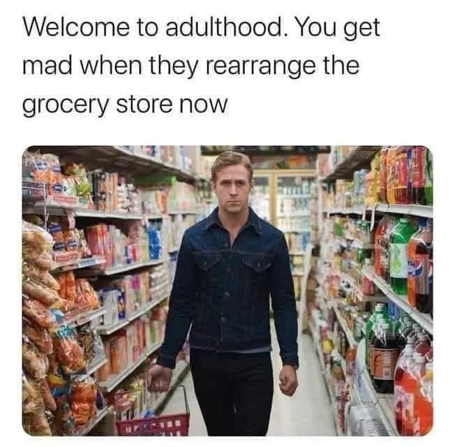 A man in a denim jacket walks down a grocery store aisle holding a shopping basket, looking intensely at the camera. The…