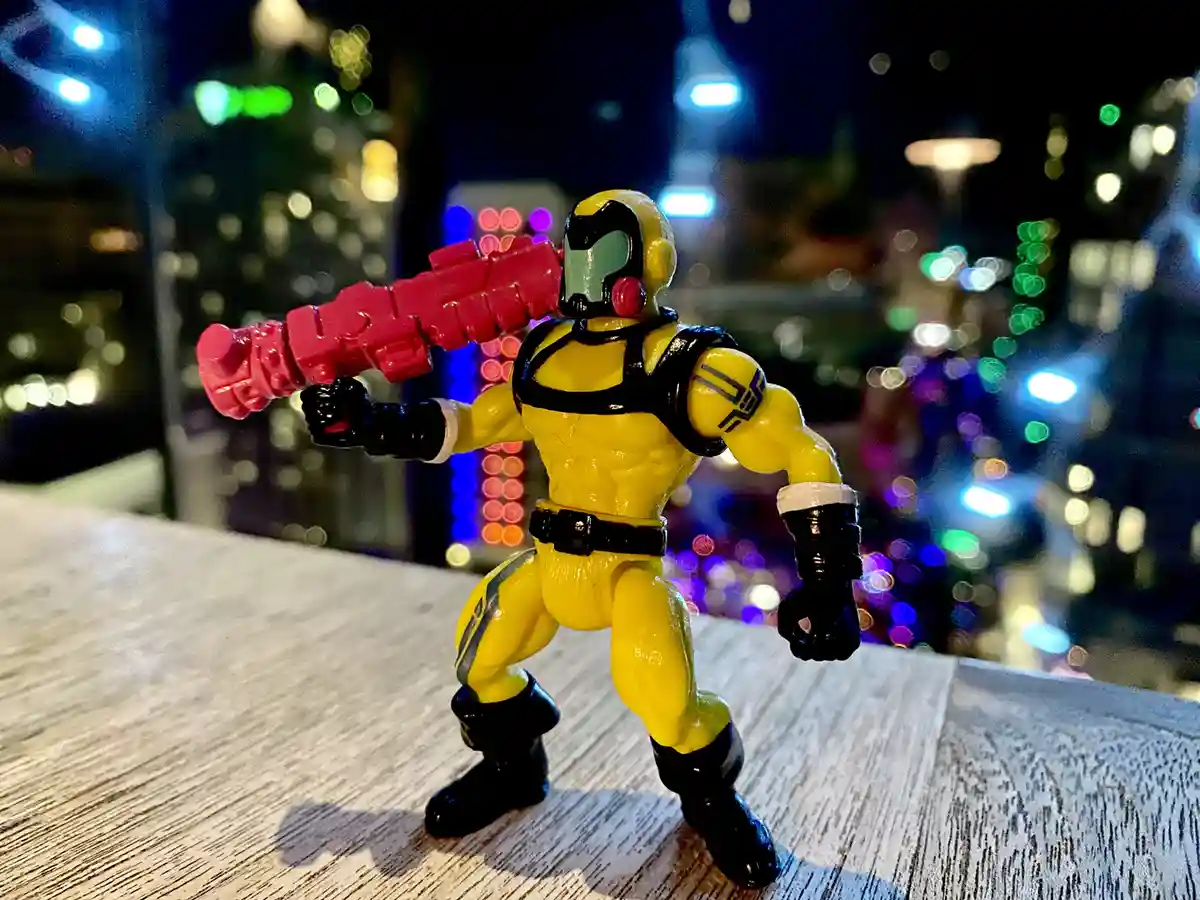 A yellow and black action figure, resembling a muscular superhero or villain with a helmet and a large pink weapon, stands…