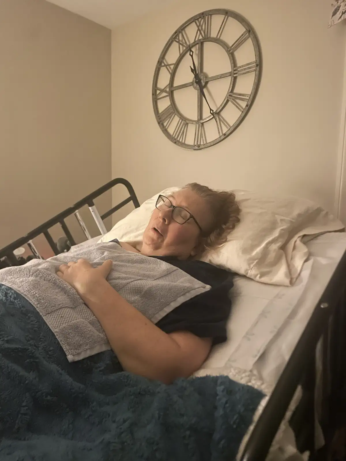 A woman with glasses lies in bed under blankets, appearing to be asleep or resting, with a large decorative wall clock…