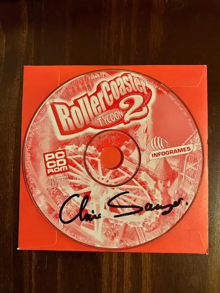A red 'RollerCoaster Tycoon 2' PC CD-ROM game disc, published by Infogrames in 2002, is shown resting on its red case. A…