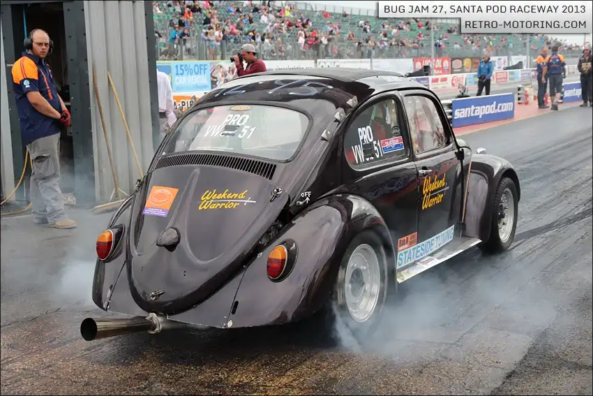 A black modified Volkswagen Beetle race car, labeled 'Weekend Warrior' and 'PRO VW-51', is on a drag strip, smoking from its…