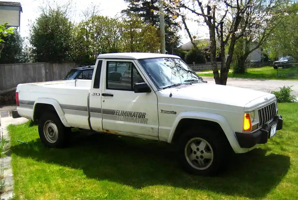 A white, rusty 1980s Jeep Comanche pickup truck is parked on a grassy lawn, showing visible wear and tear. The word…