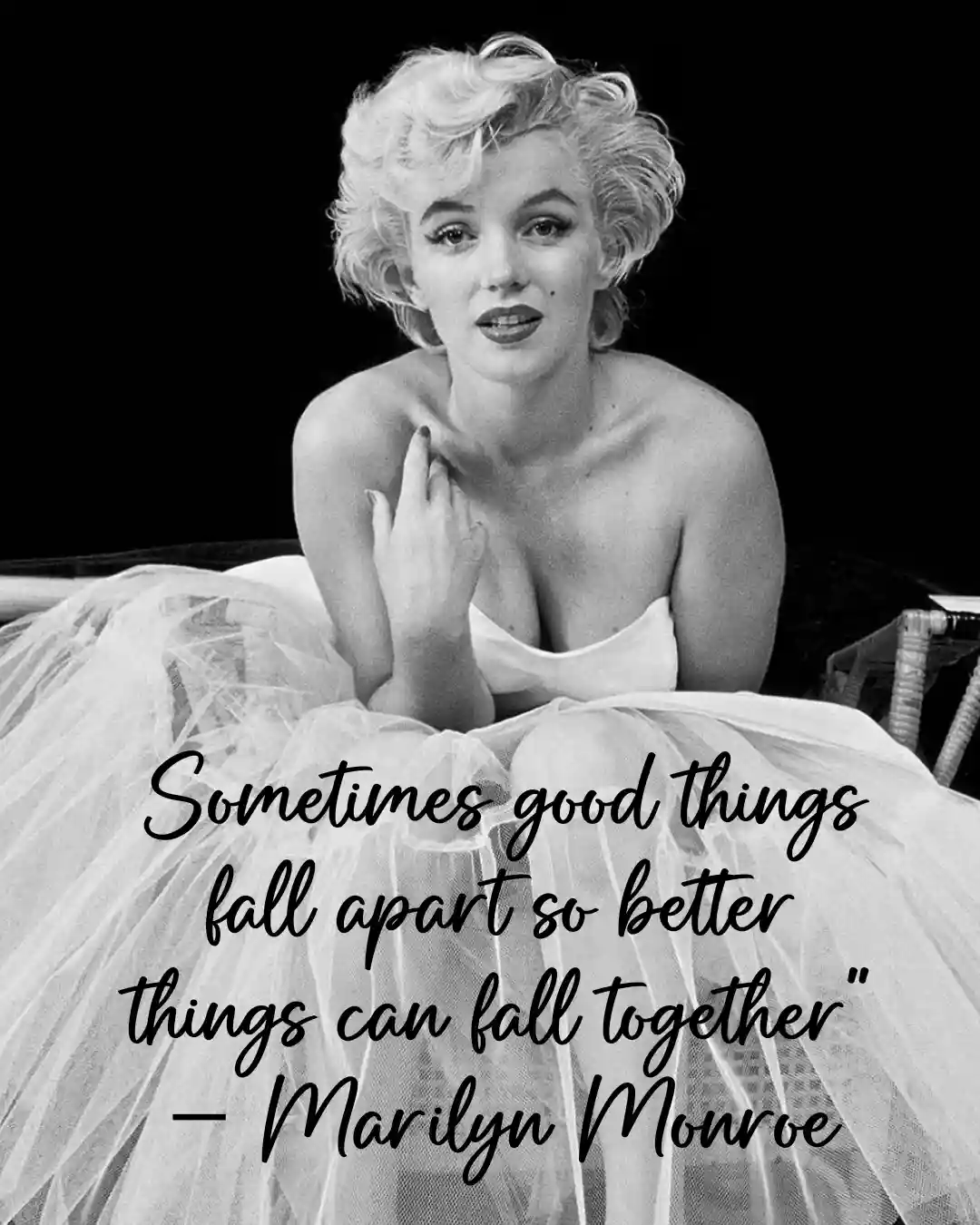 A black-and-white photo of Marilyn Monroe sitting in a white tulle dress, looking directly at the camera with her hand near…