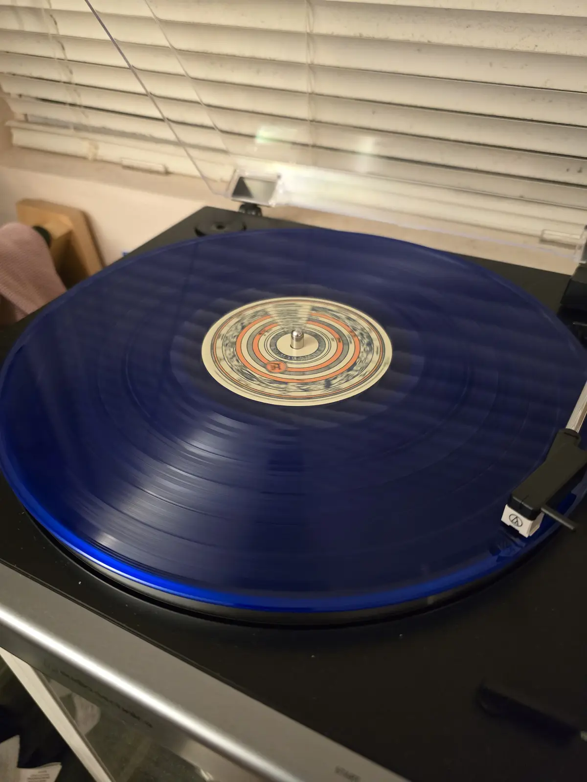 A close-up photo shows a translucent blue vinyl record spinning on a black turntable, with the tonearm positioned over the…