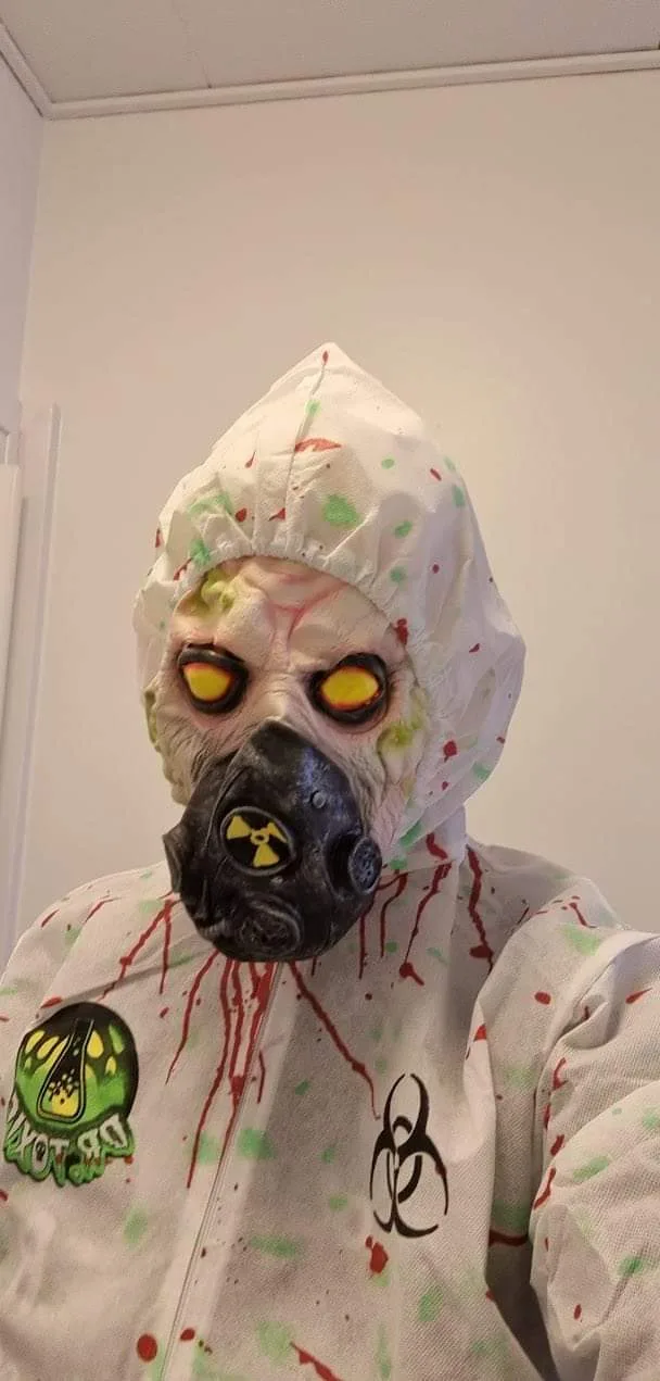 A person in a white hazmat suit with a hood and a black gas mask featuring a yellow radiation symbol, covered in red and…