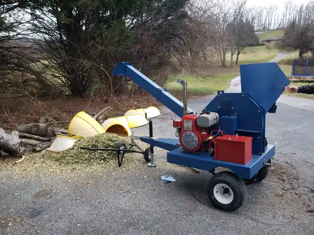 A blue and red portable wood chipper sits on a paved driveway, actively chipping wood into mulch. Several yellow plastic…