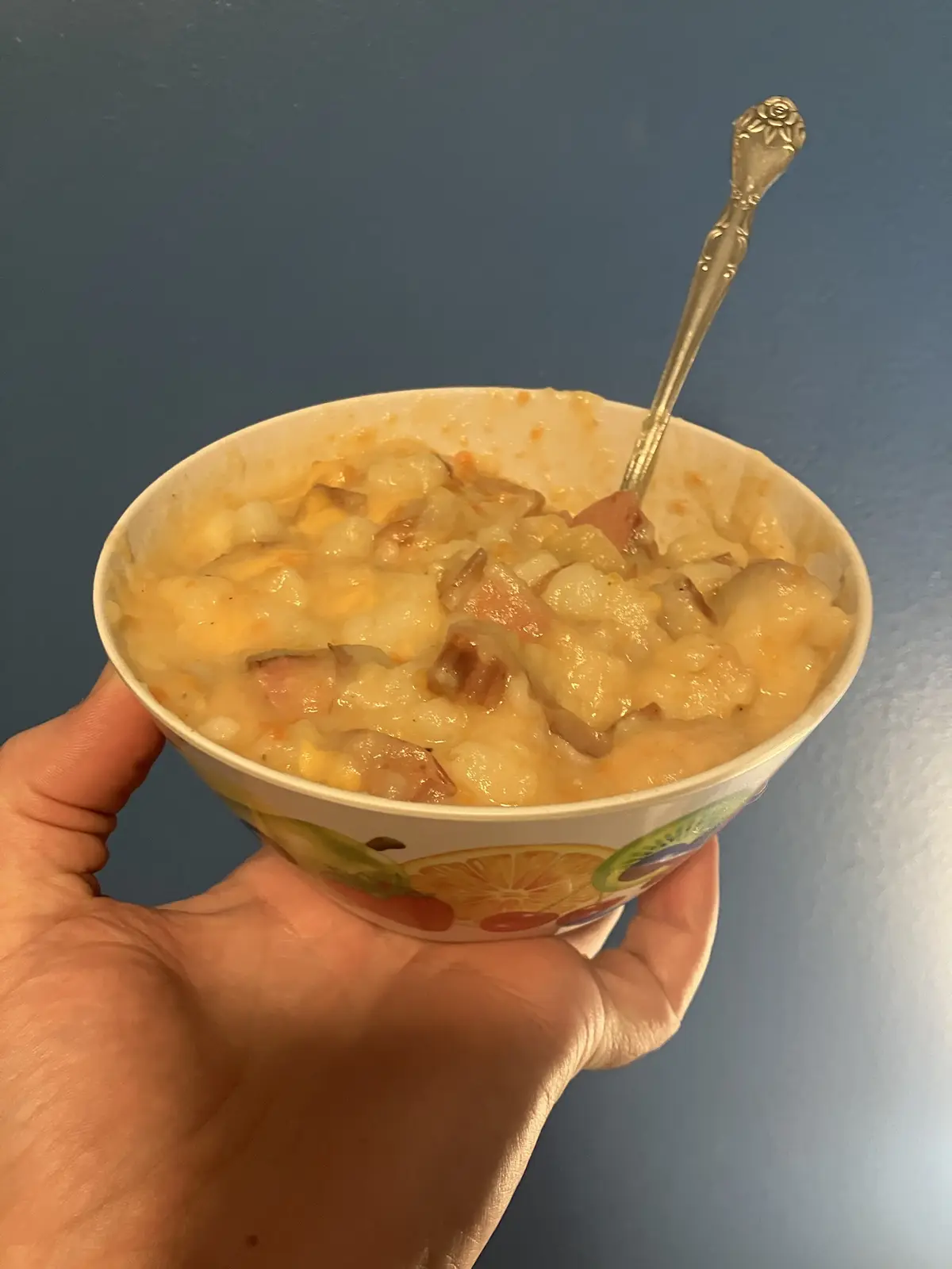 A hand holds a white paper bowl with a fruit pattern, filled with a creamy, chunky dish containing melted cheese, diced…