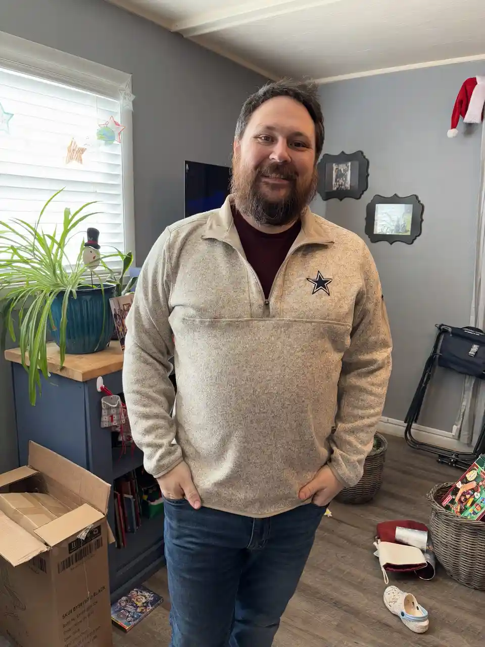 A man with a beard and a friendly smile stands in a living room, wearing a beige quarter-zip pullover with a Dallas Cowboys…