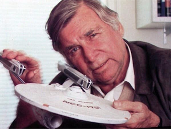 An older man with gray hair, wearing a dark suit and light shirt, holds a small model of the Starship Enterprise (NCC-1701)…