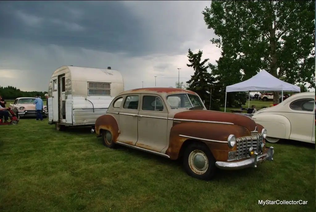 A rusty, vintage car with a faded two-tone paint job is parked on grass next to a trailer under a stormy sky. Other classic…