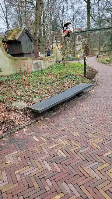 A long, dark wooden bench sits on a herringbone-patterned brick path in a park with bare trees and whimsical fairy-tale…