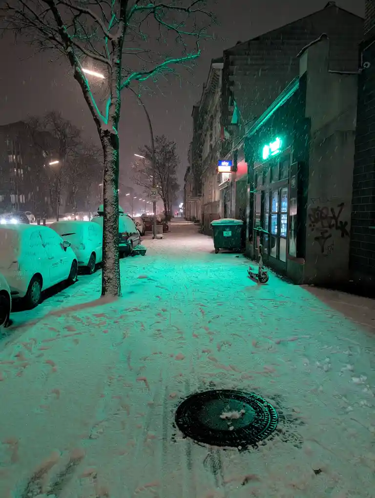 A snowy urban street at night, bathed in green light from a storefront, with snow-covered cars and a manhole cover in the…