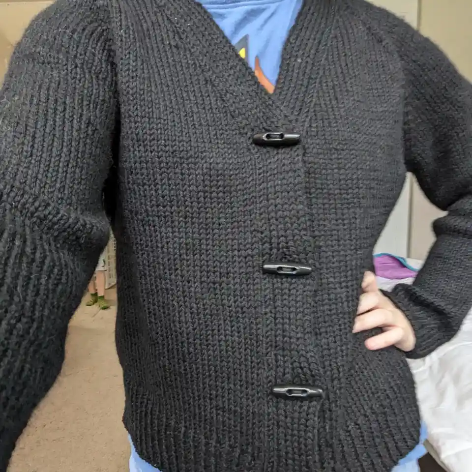 A person wearing a thick, dark gray knitted cardigan with three large black toggle buttons, standing with one hand on their…