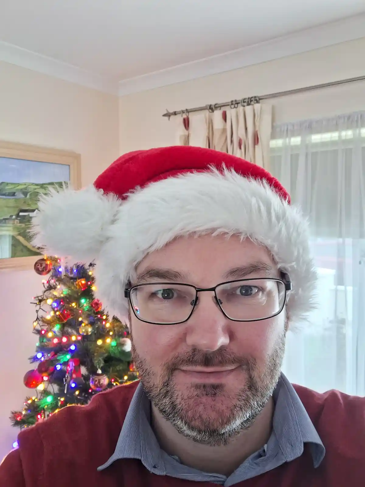 A man with glasses and a beard wears a red and white Santa hat while looking directly at the camera. Behind him is a…